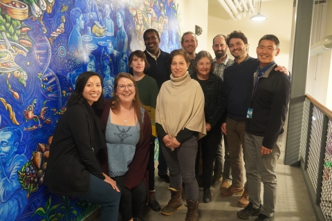 The members of City Market's Board of Directors and General Manager stand in a cluster in a hallway. On the wall to their left is a mural with a bold blue background and community food scenes.