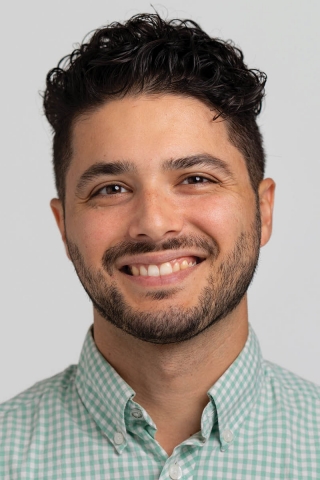 A photo of a person from the shoulders up. The person is wearing a green and white checked button down shirt. They are smiling and facing the camera. They have dark brown, curly hair.