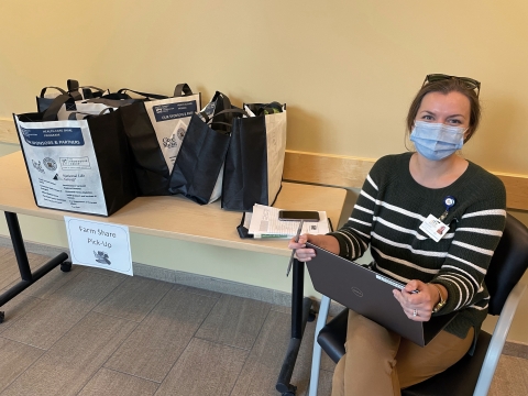 A person sits to the right of a folding table. They are wearing a blue surgical mask and have sunglasses propped on top of their head. They have a laptop on their lap and they are holding a pen. There are several reusable grocery bags on the table that are full of groceries.