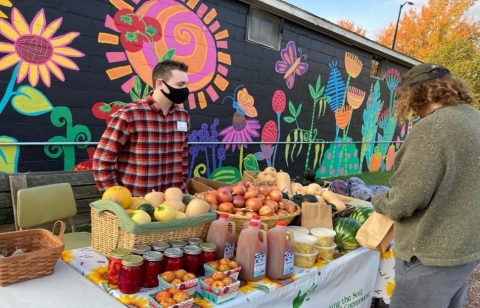 A person stands in front of a mural that is painted on the side of a building. The mural has a black background and features brightly-colored flowers and insects. The person is wearing a red plaid shirt and a black mask, and has a table in front of them that is piled high with various squash, apples, cider, and greens.