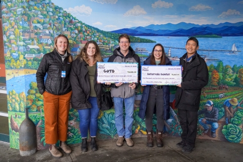Five people stand in front of a mural that depicts a hillside dotted with houses and trees sloping down to a vivid blue lake with blue mountains in the distance. The five people hold two oversized checks, one for $19,898.30 for COTS, and one for $5,167.31 for the Intervale Center.