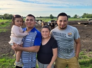 A family of farm workers stands in a field in front of a cluster of cows.