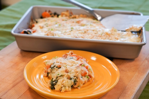 Slice of casserole with cheese, dark green kale, and sweet potato on yellow ridged plate. The plate is on a cutting board with a baking pan behind it with the rest of the casserole and spatula in the pan. The cutting board is placed on a forest green tablecloth. 