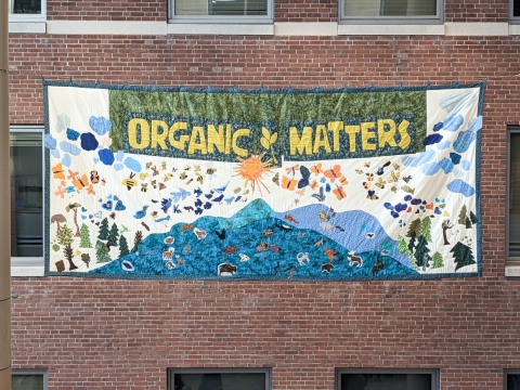 A brick wall with a quilted banner hung on it covering some windows. The banner reads Organic Matters in golden-rod colored letters on a green background. The rest of the banner is white with a blue border and teal and pale blue mountains with an orange sun. 