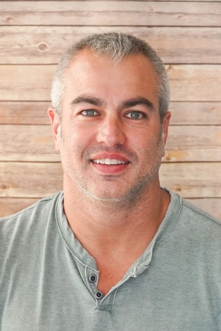 A photo of a person from the chest up in front of a wood-paneled backdrop. The person is wearing a light green polo shirt and has close-cropped silver hair.
