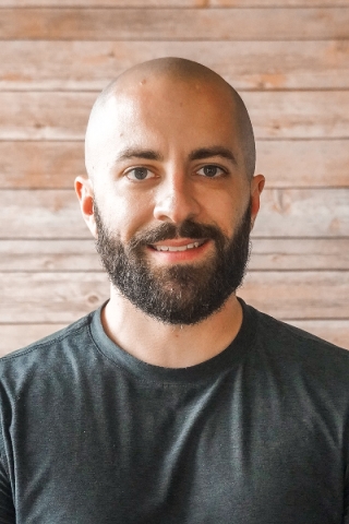 A photo of a person from the chest up. The person is wearing a plain black tee shirt. He has brown eyes and a full beard.