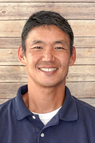A photo of a smiling person from the chest up in front of a wood-panel backdrop. The person is wearing a blue polo shirt. He has short brown hair.