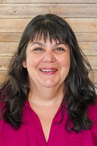 A photo of a smiling person from the chest up in front of a wood-panel backdrop. The person is wearing a pink button-down shirt. She has brown hair and brown eyes.