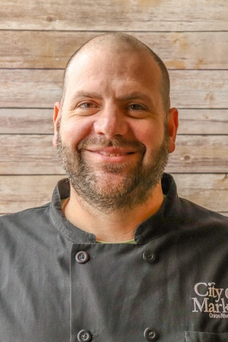 A photo of a smiling person from the chest up in front of a wood-panel backdrop. The person is wearing a black chef's coat. he has a close-cropped beard.