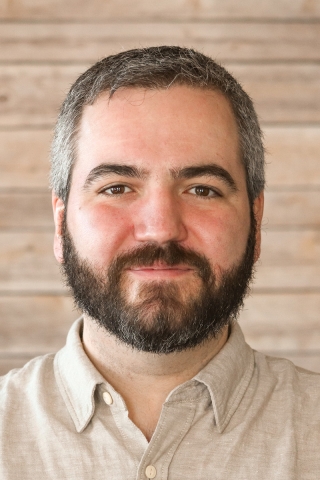 A photo of a smiling person from the chest up in front of a wood-panel backdrop. The person is wearing a khaki polo shirt. He has short brown hair and a short brown beard..