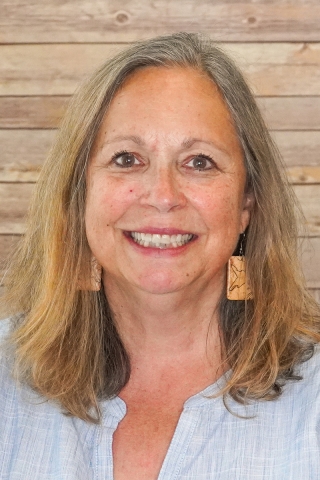 A photo of a person from the shoulders up in front of a wood-panel backdrop. The person is wearing a light blue shirt. She has light brown shoulder length hair and wears large square earrings.