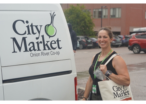 A smiling woman stands to the right of a van that has the City Market logo on the side. She is wearing a black shirt and has a City Market tote bag filled with groceries over her shoulder.