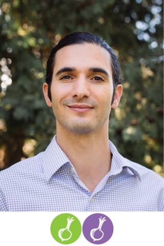 A photo of a person from the chest up. The person is wearing a light blue, crisp button-down shirt. They have black hair. They are standing in front of blurry dark green foliage.