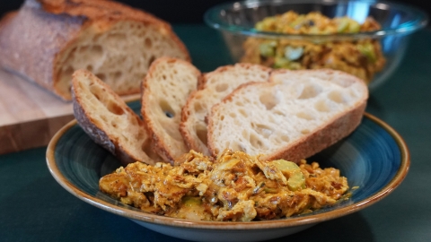 Our curry chicken salad is on a plate with four slices of hearty bread piled behind it. Behind that plate to the right is a bowl of chicken salad, to the left a cutting board with the rest of the loaf. 