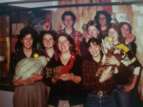 A group of eight women and one man pose for a photo with a variety of produce items, including tomatoes, apples, corn, an artichoke, a pineapple and a very large gourd.