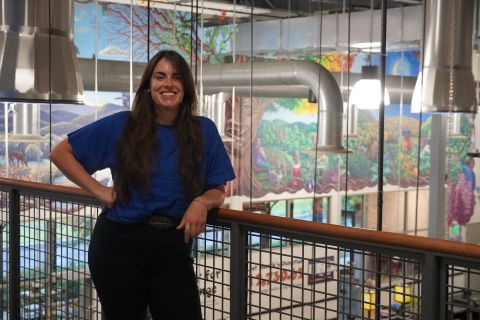 A person stands leaning against a railing of a mezzanine overlooking the South End City Market store. She is wearing a blue shirt and black pants. She has long dark hair and is smiling. Behind her, lights and piping hang from the ceiling. Behind the lights, there is a mural.
