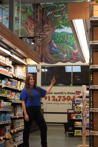 A person stands in the wellness aisle of the South End City Market store. She is pointing to a mural above the check-out area that depicts a woman sitting under a tree. The woman and the person in the photo look very similar to one another.