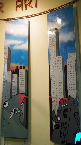 Two 6' painted wooden planks. There is a blue sky with clouds in the background as well as tall buildings. In the front is a shark on the left plank, and a robot on the right plank. The robot is directing lasers from its eyes to the sharks nose.
