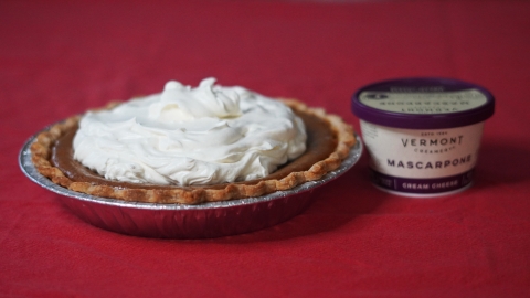 A pumpkin pie with white whipped topping on a red background. Beside the pie there is a container of Vermont Creamery Mascarpone.