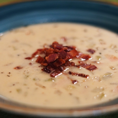 A close up of the soup in a teal bowl. The soup is white, with bits of celery, carrot, and onion floating within. Topping the center of the soup is a pile of crumbled bacon. 