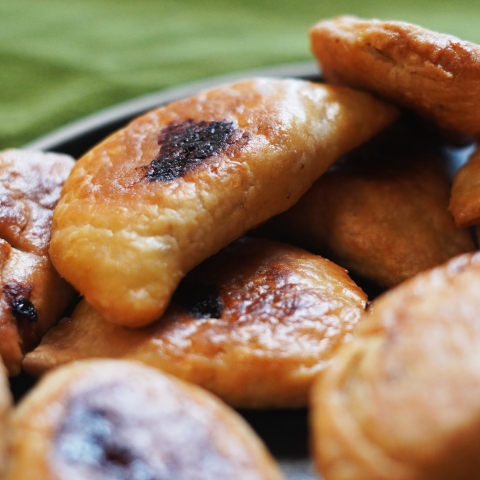 A close up of many turnovers. Many are in soft focus in the foreground of the image, and 2 are in clear focus with bits of browned cheese poking out from the center of the pastry. 