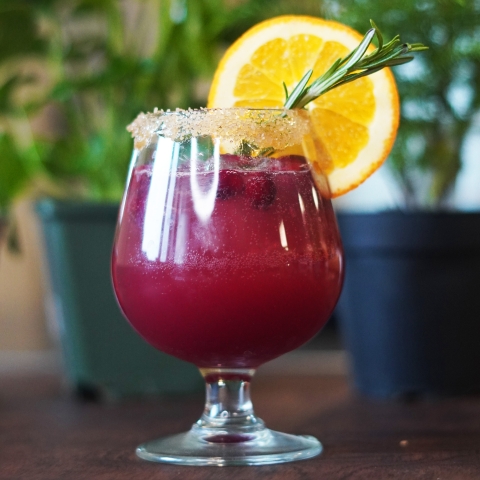 The sunset in a little cocktail glass, there is an orange slice on the rim and a sprig of rosemary sticking out of the glass.