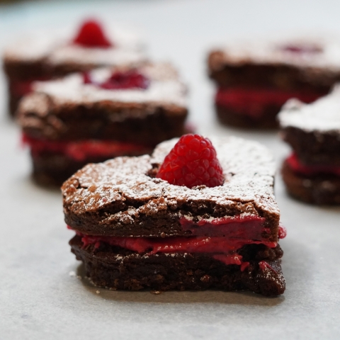 A close up of a heart shaped brownie, with a layer of red cream in the middle, and topped with a raspberry. behind it, and out of focus, are more brownies!