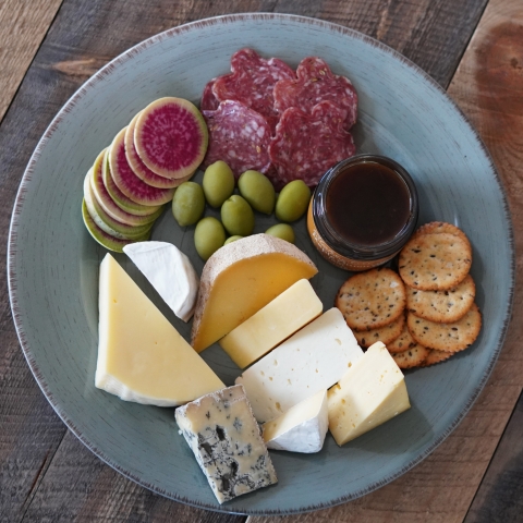 A plate of cheeses with classic cheese board accoutrements like salumi, maple fig jam, crackers, olives and daikon.