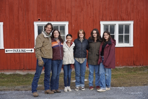 A family of six - dad, mom, and four daughters - stands in front of a bright red barn. 