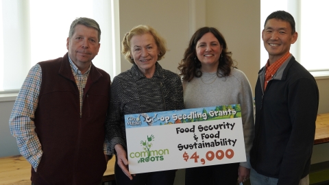 A group of people gathered around a sign that says "Common Roots - Food Security & Food Sustainability - $4,000"