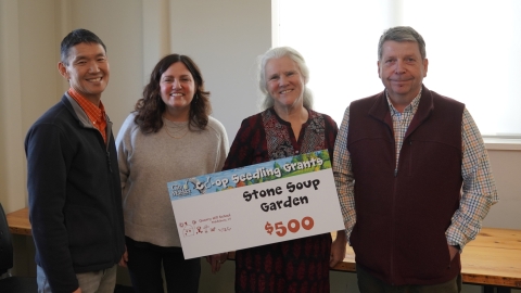 A group of people gathered around a sign that says "Quarry Hill School - Stone Soup Garden - $500"