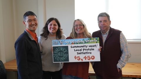 A group of people gathered around a sign that says "Hardwick Area Food Pantry - Community Meat / Local Protein Initiative - $5,450"
