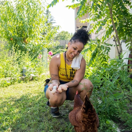 A person crouches in a green yard with tall greenery growing all around her. She holds a handful of fresh eggs and smiles in camaraderie at two chickens. She has dark hair piled in a large bun on top of her head. a