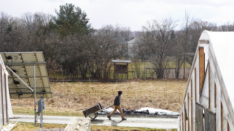 A person pulls a cart along a walking path through a farm. He passes solar panels and a greenhouse. A light snow dusts the field and fills the air.