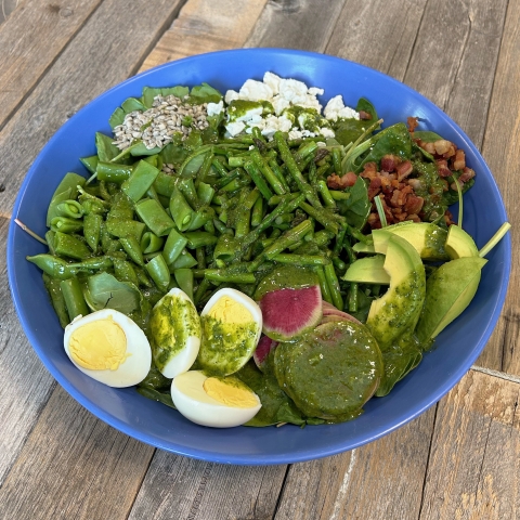 A salad in a blue bowl. Greens are topped with sunflower seeds, watermelon radish, boiled eggs, pancetta, asparagus, goat cheese, avocado, and snap peas