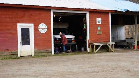 A red barn with a Pomykala Farm sign on the side. There's an open garage door, and through it, a white van is visible. A person in a red coat is working on the front of the van.