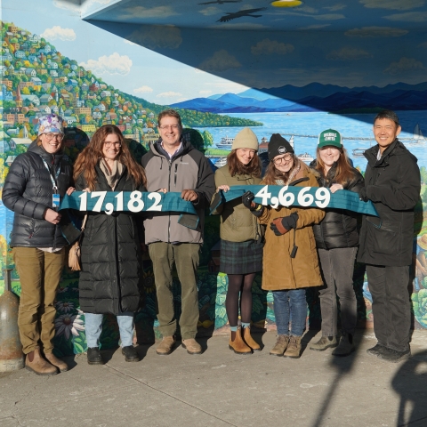 Seven people in winter coats stand in front of the mural outside the Downtown City Market store. They are holding green banners with dollar totals - $17,182 and $4,669.