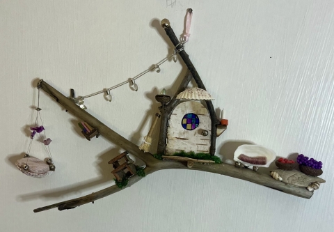 A fairy house situated on a twig. A pink hanging chair on the far left with birds on the wire. There's string lights across the top, below that is a little door with a mini broomstick next to it. On the right end of the branch is a chair made from a shell and two baskets resembling fruit.