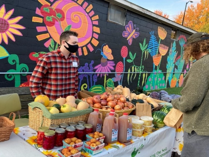 A person stands in front of a mural that is painted on the side of a building. The mural has a black background and features brightly-colored flowers and insects. The person is wearing a red plaid shirt and a black mask, and has a table in front of them that is piled high with various squash, apples, cider, and greens.