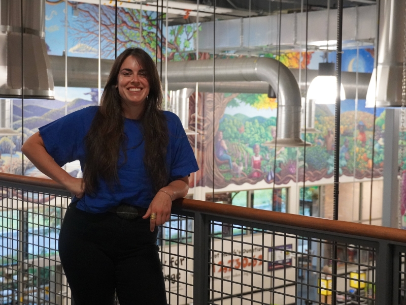 A person stands leaning against a railing of a mezzanine overlooking the South End City Market store. She is wearing a blue shirt and black pants. She has long dark hair and is smiling. Behind her, lights and piping hang from the ceiling. Behind the lights, there is a mural.