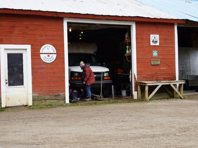 A red barn with a Pomykala Farm sign on the side. There's an open garage door, and through it, a white van is visible. A person in a red coat is working on the front of the van.